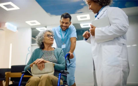 Técnico en enfermería acompañando a adulta mayor en silla de ruedas junto a profesional de la salud en centro médico.