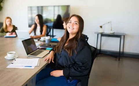 Profesional en administración pública sonriendo frente a su computador durante reunión de equipo.