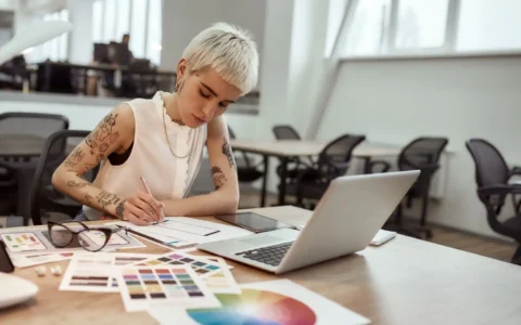 Diseñadora gráfica con tatuajes realiza boceto con lápiz entre paletas de colores y computador portátil sobre mesa de oficina creativa.