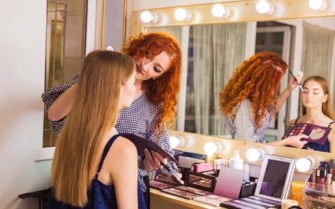 Esteticista aplica maquillaje con brocha a clienta sentada frente a tocador iluminado lleno de cosméticos y pinceles.