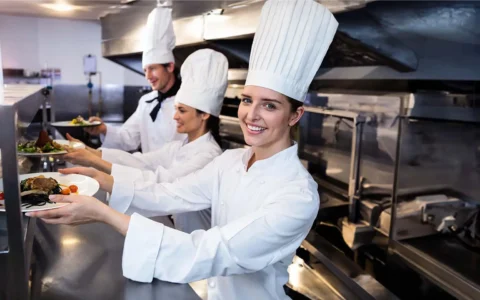 Chefs con gorros blancos entregan platos gourmet en cocina industrial equipada, destacando formación en gastronomía internacional.