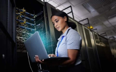 Ingeniera revisa líneas de código proyectadas sobre laptop en sala de servidores con cables y racks de redes empresariales.