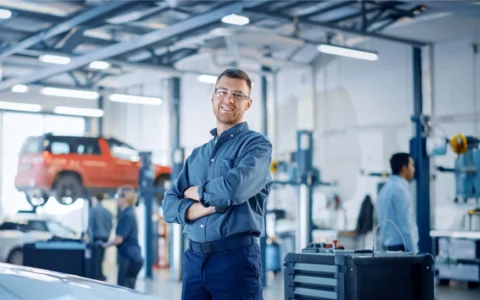 Ingeniero automotriz con lentes y vestido de azul marino cruza los brazos en taller con vehículos elevados y herramientas tecnológicas modernas.