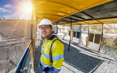 Ingeniero en minas con casco y ropa reflectante sonríe junto a rajo a cielo abierto iluminado por el sol y estructura metálica.