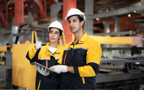 Ingenieros en prevención de riesgos con casco y uniforme revisando condiciones de seguridad en planta industrial con una tablet.