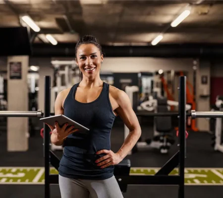 Entrenadora personal en gimnasio con pesas, sostiene tablet y sonríe lista para guiar rutina física personalizada a usuarios.
