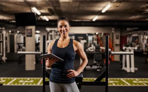 Entrenadora personal en gimnasio con pesas, sostiene tablet y sonríe lista para guiar rutina física personalizada a usuarios.