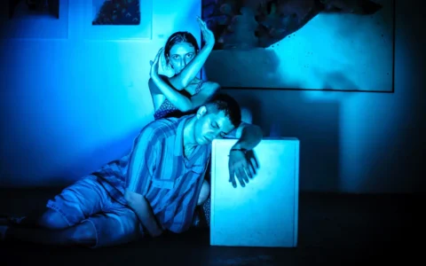 Pareja de actores en escena teatral bajo luz azul intensa, interpretan gesto expresivo sobre cubo blanco en escenario oscuro.