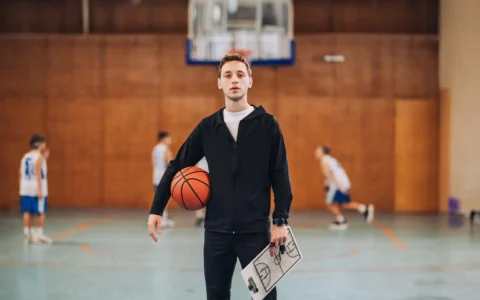 Entrenador deportivo sostiene balón y pizarra táctica en gimnasio con niños practicando baloncesto en segundo plano.