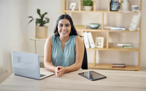 Técnica en administración de empresas sonriendo frente a su computador en oficina con estantería al fondo.