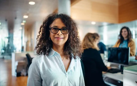 Mujer profesional en administración pública sonriendo en un entorno profesional con equipo de trabajo al fondo.
