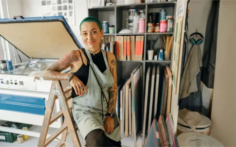 Artista con delantal y cabello teñido posa sonriente en taller de serigrafía lleno de tintas, marcos y afiches.
