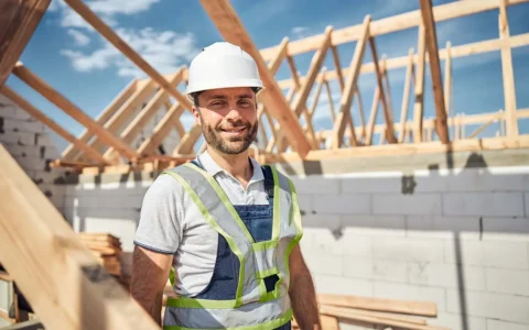 Obrero técnico en construcción con casco blanco y arnés de seguridad posa frente a obra en madera bajo cielo despejado.