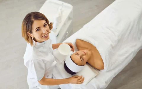 Cosmetóloga sonriente aplica tratamiento facial a paciente recostada en camilla, con productos y ambiente de estética limpio.