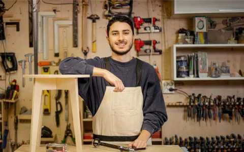 Diseñador en taller de carpintería apoya brazo sobre mesa mientras sonríe junto a mueble de madera y herramientas organizadas.