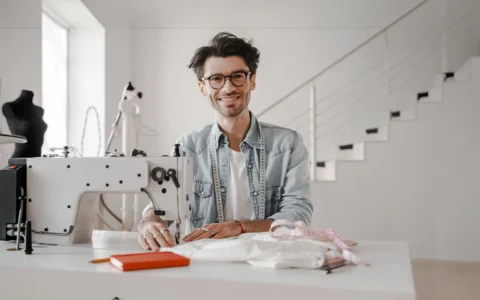 Diseñador frente a máquina de coser, con cinta métrica y telas, rodeado de libretas, maniquíes y elementos de confección.