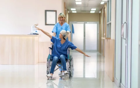 Técnico en enfermería oncológica empujando a paciente en silla de ruedas en pasillo de hospital.