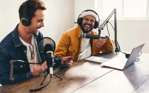 Dos locutores sonrientes con auriculares graban programa radial con micrófonos, laptop y fondo de estudio acústico moderno.