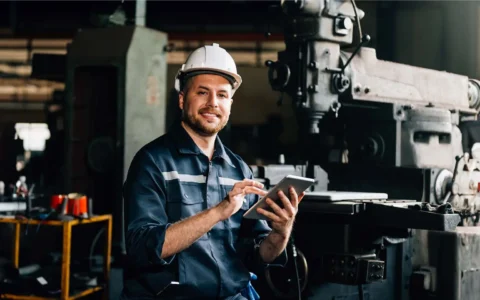 Técnico en mantenimiento industrial sonriente con casco y tablet inspecciona torno mecánico en planta de mantenimiento con luz tenue.
