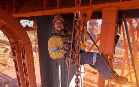 Técnico minero con arnés y casco trabaja suspendido con cadenas en estructura metálica de faena minera a cielo abierto.