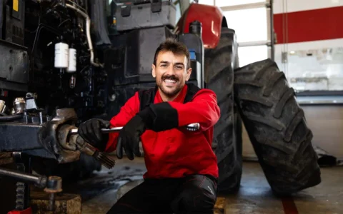 Mecánico con overol rojo y guantes ajusta pieza en rueda de maquinaria pesada, sonriendo en taller con neumáticos grandes.