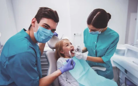 Técnicos dentales revisan dientes de niña en consulta mientras uno sonríe y el otro utiliza herramientas de exploración dental.