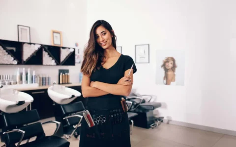 Estilista joven con herramientas en la cintura posa sonriendo en moderno salón de peluquería con lavacabezas y estanterías.