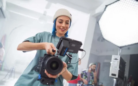 Camarógrafa sonriente ajusta cámara profesional en estudio de filmación con luz LED, trípode, monitor y fondo desenfocado.