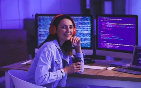 Técnica en programación y análisis de sistemas trabajando frente a múltiples monitores con código, usando audífonos y sosteniendo una taza.