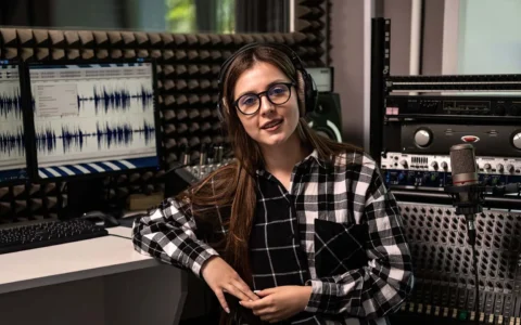 Estudiante de sonido con audífonos opera consola digital frente a pantallas con forma de onda y paneles acústicos grises.