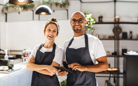 Pareja de empresarios gastronómicos sonrientes en un café, usando delantales, uno con una tablet y el otro cruzando los brazos.