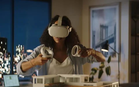 Estudiante usa visor de realidad virtual y controles sobre maqueta arquitectónica en oficina moderna con iluminación nocturna.