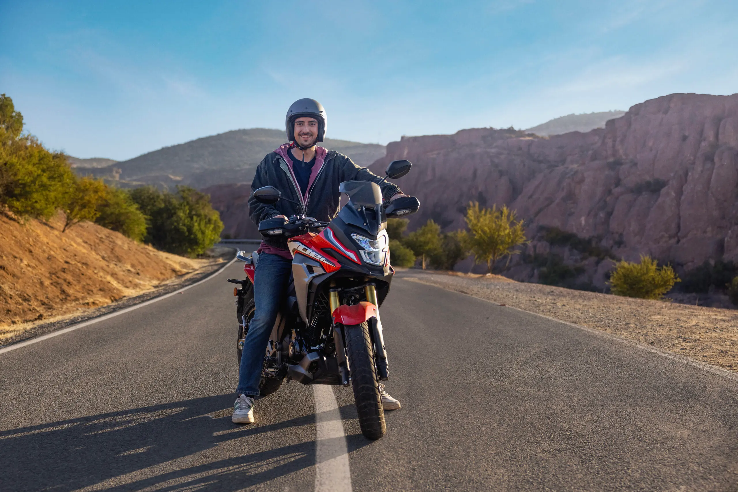 Hombre con casco sobre moto roja detenido en carretera rural entre cerros, día despejado.