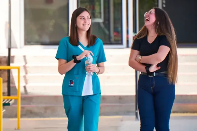 Dos estudiantes conversan y ríen mientras caminan por el campus de AIEP, una de ellas con uniforme clínico y botella de agua en la mano.